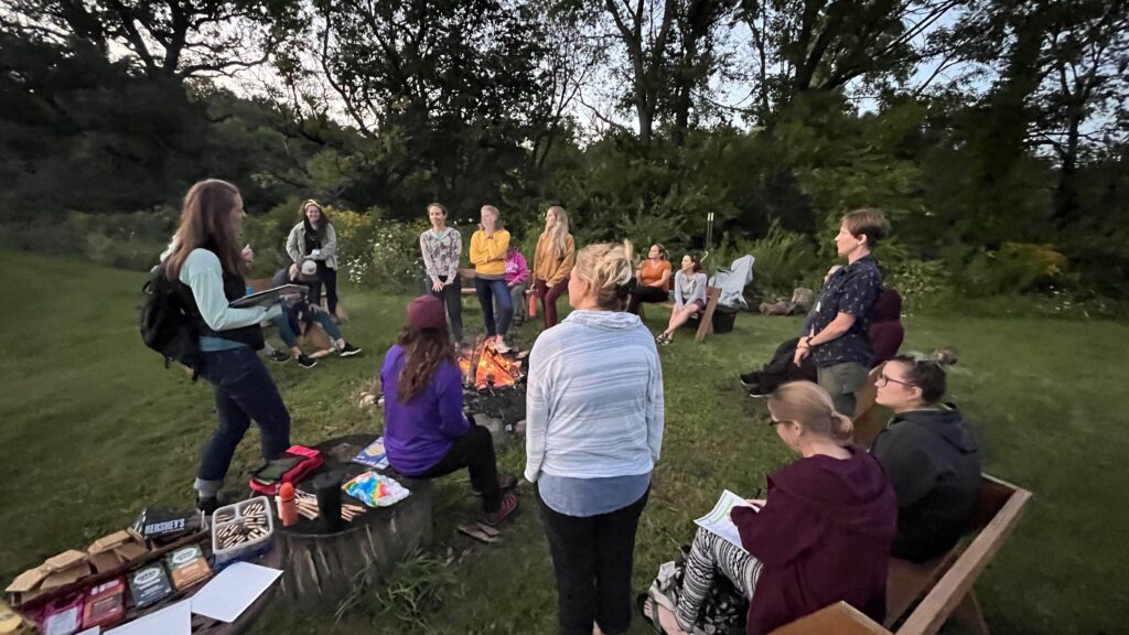 retreat attendees sitting and standing around a fire pit with a s'mores station nearby